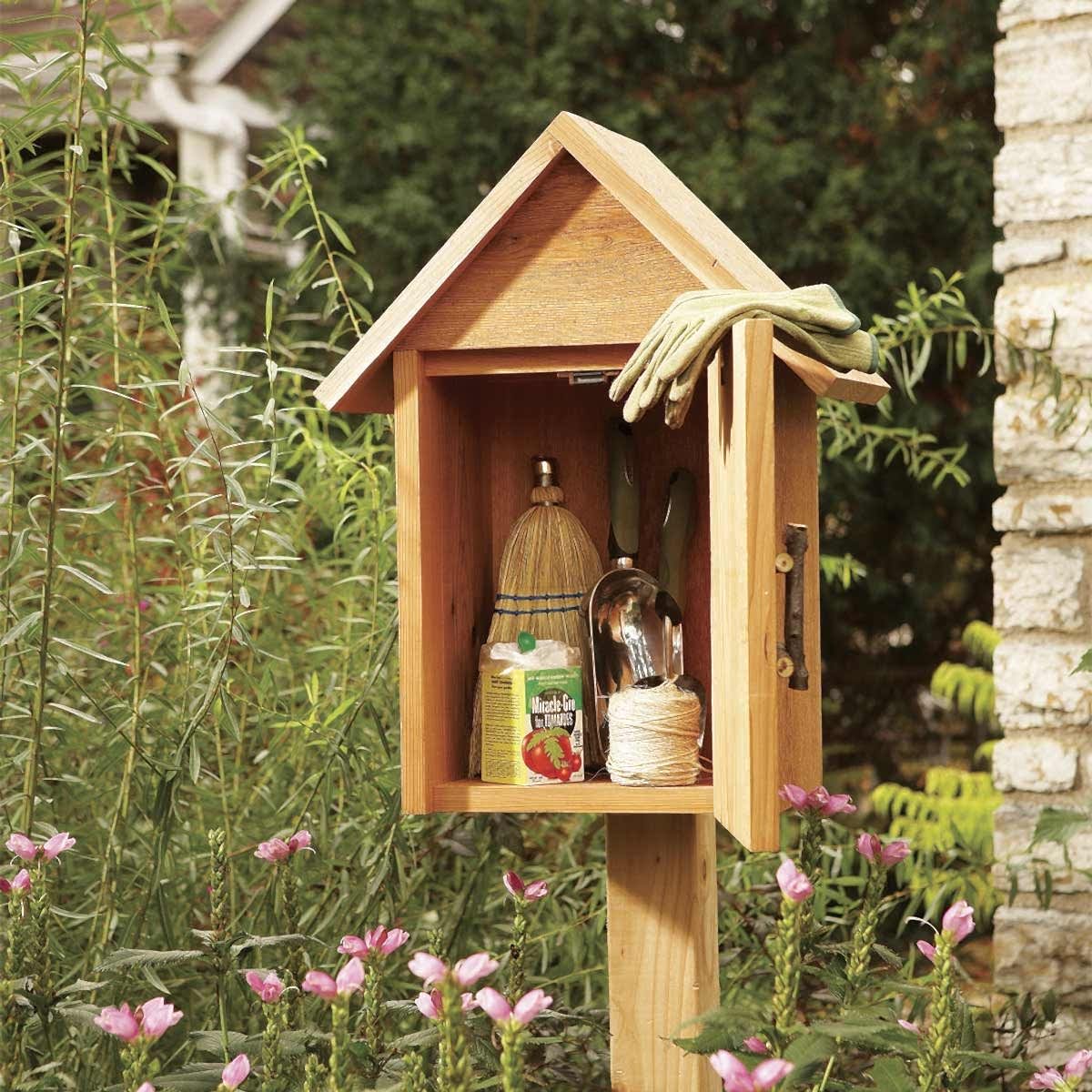 A garden shed stands on a post, displaying gardening tools inside. Pink flowers grow nearby, with greenery providing a natural backdrop. A pair of gloves rests on top.