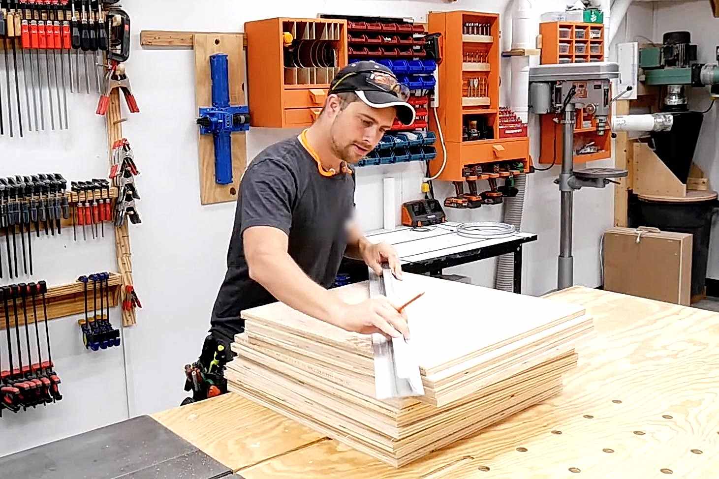 A man is measuring a stack of plywood sheets on a workbench in a well-equipped workshop filled with tools and materials.