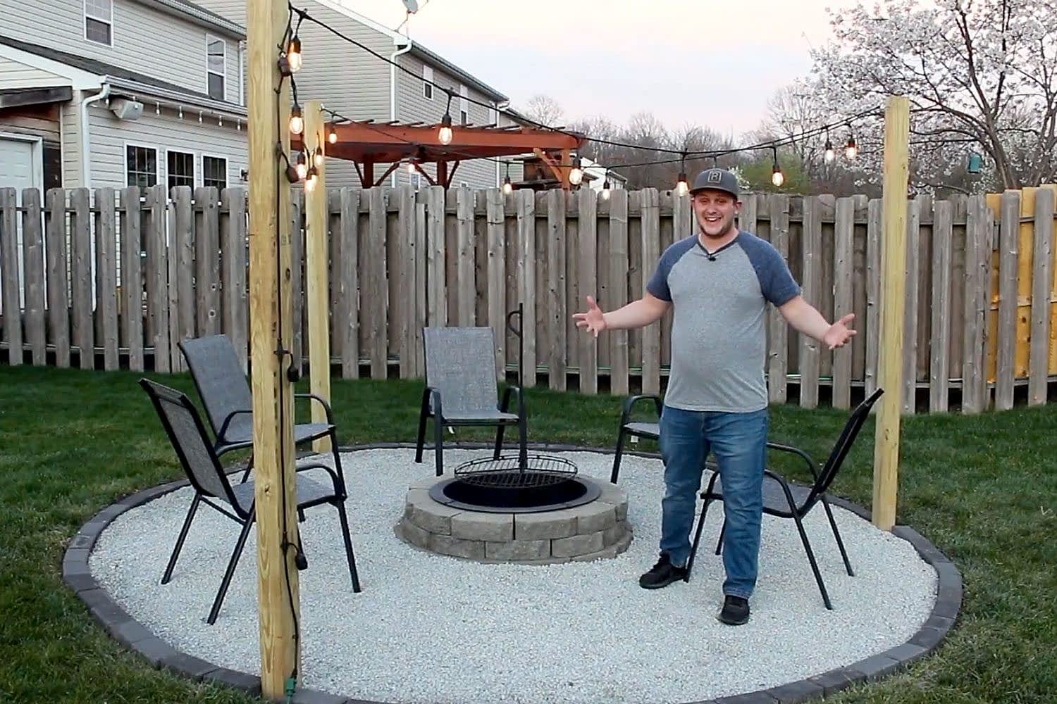 A man stands by a fire pit surrounded by chairs on a gravel circle, illuminated by string lights in a backyard setting.